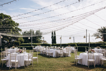 Wedding banquet. Tables in a green meadow are decorated with flower arrangements, on the tables are white tablecloths, plates with napkins, glasses and candles, cutlery. White bulbs hang over tables