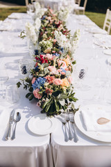 Wedding banquet. Tables in a green meadow are decorated with flower arrangements, on the tables are white tablecloths, plates with napkins, glasses and candles, cutlery. White bulbs hang over tables