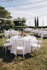 Wedding banquet. Tables in a green meadow are decorated with flower arrangements, on the tables are white tablecloths, plates with napkins, glasses and candles, cutlery. White bulbs hang over tables
