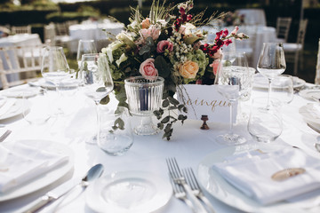 Wedding banquet. Tables in a green meadow are decorated with flower arrangements, on the tables are white tablecloths, plates with napkins, glasses and candles, cutlery. White bulbs hang over tables