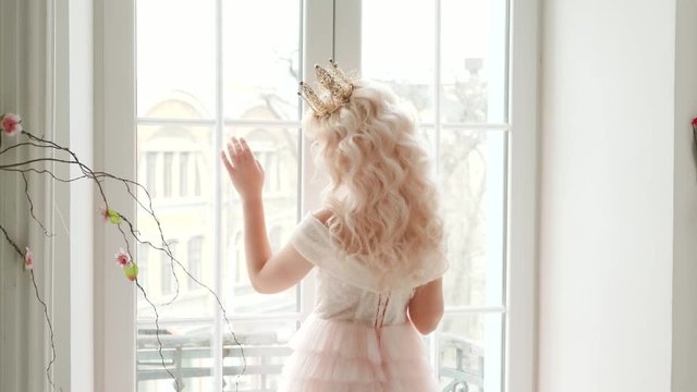 young beautiful woman medieval queen. Hairstyle blonde long wavy hair crown. Sad Girl Princess turned away stay at home touching window concept hope. Vintage style room lush pink dressr rear back view