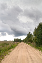 Dirt road between forest and field under stormy cloudy sky