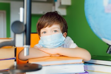 Tired boy studying at home in a protective face mask during CoVid-19 quarantine, distance learning online with a laptop, a child doing homework for school. Schoolboy stayed at home.