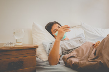 Asian man lying in bed wearing protective mask measuring temperature.