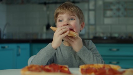 Young boy eating pizza alone at home on blue kitchen. Close up of one kid enjoying big pizza in slow motion. 