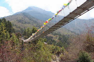 One of many pedestrian suspension bridges with waving prayer flags in Himalayas on the way to Everest Base Camp in Nepal. Mountainside covered with forest. Clouds on the sky. - obrazy, fototapety, plakaty