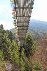 One of many pedestrian suspension bridges with waving prayer flags in Himalayas on the way to Everest Base Camp in Nepal. Bottom up view. Mountainside covered with forest. Theme of travel in Nepal. - obrazy, fototapety, plakaty