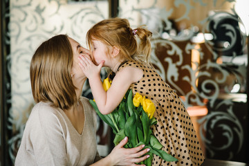 Cute little girl greeting mother and gives her a bouquet of flowers tulips at home. Mother's day...