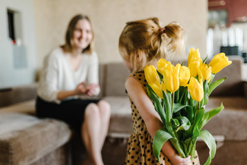 Cute little girl greeting mother and gives her a bouquet of flowers tulips at home. Mother's day...