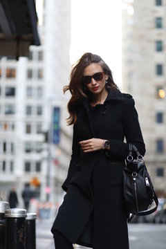 A woman in total black is walking down in New York street. Black coat, bag, jeans, boots, sunglasses