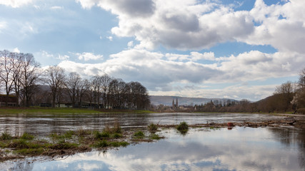 The big river is called Weser. In the background you can see the city of Höxter with the church with two towers. The river has burst its banks. Clouds move across the city in the blue sky.