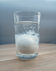 Glass cup with mineral water and an egg inside. The egg is dipped inside the glass and is enveloped in a white consistency. Close-up