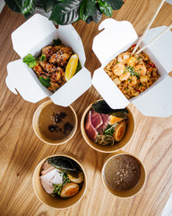 Top view on variety of japanese delivery food in brown boxes with ramen, rice, fried chicken and chopsticks on the wooden table, vertical