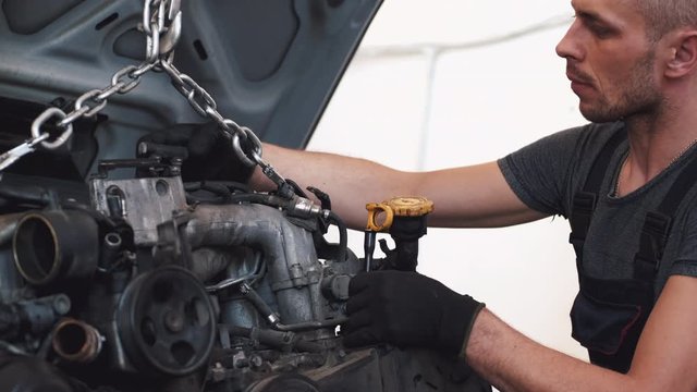 Man wears uniform and gloves. He puts motor in car. Mechanic repairs machine in garage.
