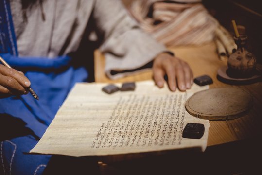 Closeup of a person writing in Hebrew on an aged paper on the table under the lights