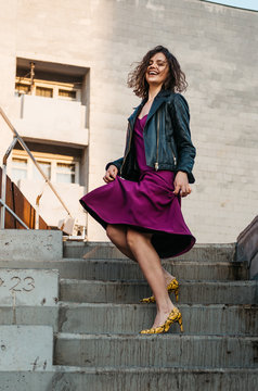Young European female with short curly hairy in a classy long magenta dress black jacket and yellow heels walking down stairs laughing