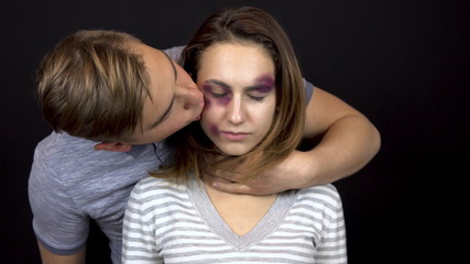 A young man strokes and kisses a young woman. A woman with bruises on her face. Quarrel in a young family. Domestic violence. On a black background