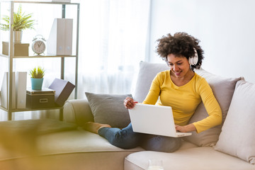 Young beautiful african american woman relaxing and listening to music using headphones. Smiling young woman watching a video with her laptop while siting on a sofa