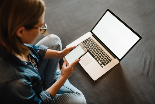 Girl checking social apps on white blank screen smartphone in home. Woman working and using laptop computer in the bedroom. Freelancer. Writing, typing. Communication and technology concept. Top view.
