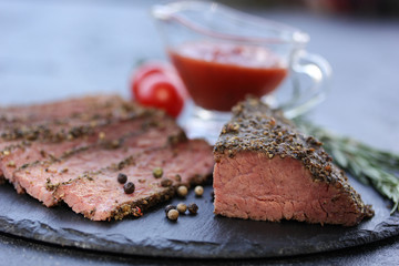 Grilled beef pastrami with rosemary, spices, cherry tomatoes and ketchup in a bowl on a black board on a dark background. Fried meat. Background image, copy space