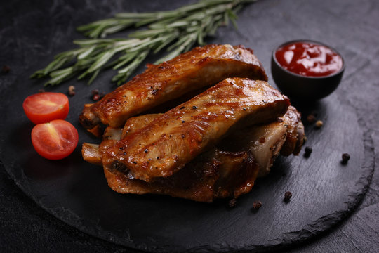 Fried pork ribs with a fried crust with spices, rosemary, cherry tomatoes and ketchup in bowl on a black board on a black background. Meat on grill. Background image, copy space