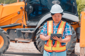 Woman Engineering wearing a white safety helmet standing In front of the backhoe And are using tablet to check the blueprint with construction