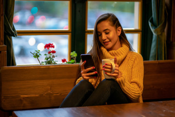 Woman sitting on sofa in front of window, drinking coffee, surfing internet on mobile phone