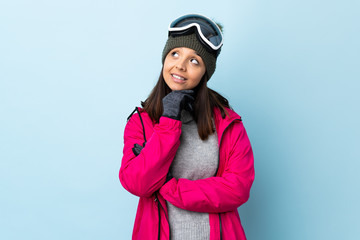 Mixed race skier girl with snowboarding glasses over isolated blue background looking up while smiling.