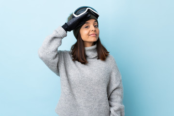 Mixed race skier girl with snowboarding glasses over isolated blue background saluting with hand with happy expression.