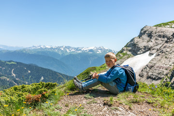 Naklejka na ściany i meble A boy traveler with hiking poles and a backpack is resting in the summer on top of a mountain with snow.