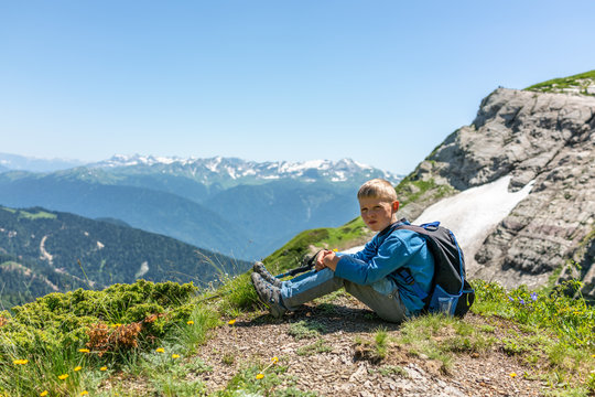 A boy traveler with hiking poles and a backpack is resting in the summer on top of a mountain with snow. © Dmitrii Potashkin