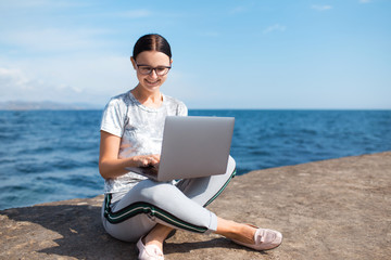 Happy young woman sitting with a laptop by the sea. Webinar online teaching concept. Online work.