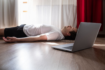 Young man with laptop meditating on floor and lying in Shavasana at living room.