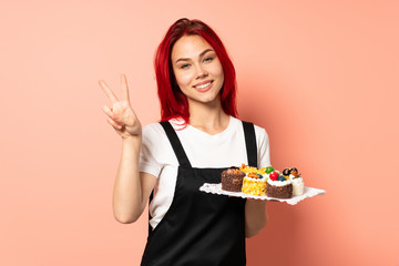 Pastry chef holding a muffins isolated on pink background showing victory sign with both hands