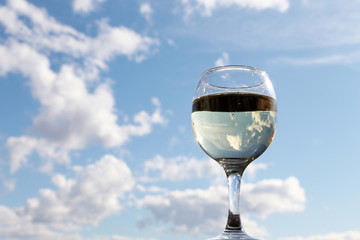 Glass of white wine on background of blue sky, clouds are reflected in alcohol drink. Concept of celebration, home party, romantic dinner outdoors