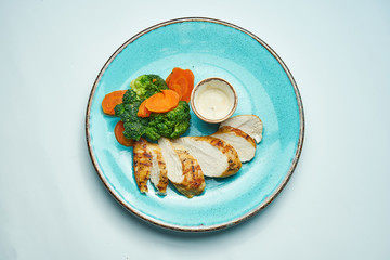 Healthy, dietary food - baked chicken fillet with boiled carrots and broccoli in a blue ceramic plate isolated light grey background.