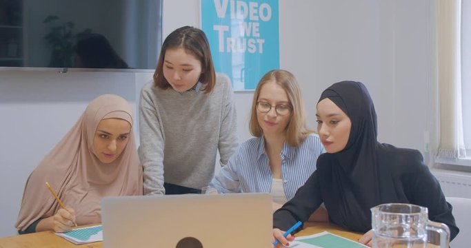 Four confident cheerful diverse colleagues office women hijab discussion business plan use laptop laughter joy