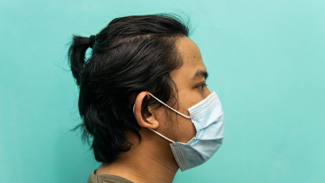 The Side View Portrait Of A Young Malay Man Wearing 3 Layer Surgical Face Mask On Isolated Blue Background.