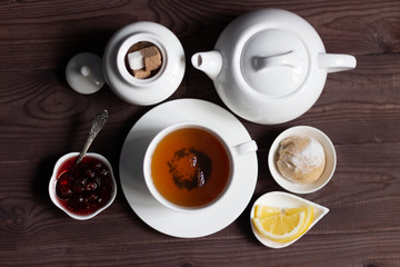 Flat lay on cup of herbal tea, teapot, sweet bun, sugar pot with cap, raspberry jam, lemon on brown wooden background