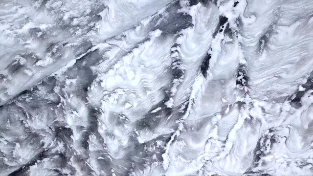 Aerial top down footage of ice glacier next to the lake with snow in the air. Glacier creates abstract textures, snow is snowing as the drone goes up. 