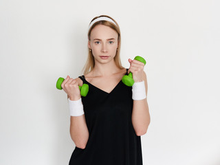 Fototapeta na wymiar Beautiful young sporty woman in black outfit doing exercises with green dumbbells to stay healthy and active on self isolation in home quarantine posing against white wall