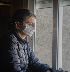 woman looking out the window while wearing a protective mask on her face during the virus epidemic.