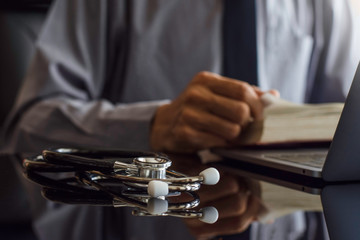 Male doctor with grey shirt reading text book, research data and working on laptop computer with medical stethoscope on the desk in room at clinic. Online learning and education concept.