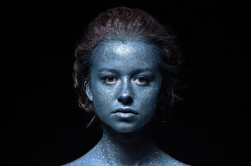 girl with a blue face in sparkles close-up against a black wall looks at the camera