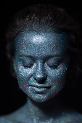 girl with a blue face in sparkles close-up against a black wall 