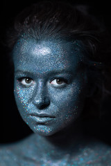 girl with a blue face in sparkles close-up against a black wall looks at the camera