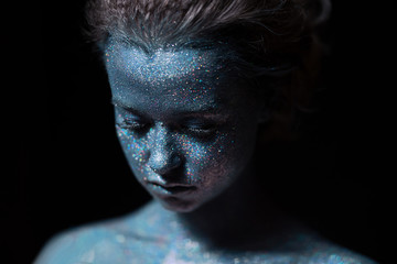 a girl with a blue face in profile in sparkles close-up against a black wall looks forward