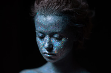 a girl with a blue face in profile in sparkles close-up against a black wall looks forward