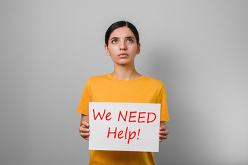 we need help. dramatic portrait of a young woman in yellow t-shirt holding a placard and looking up asking for help against grey background