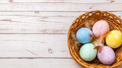 top view of easter eggs hand painted in pastel colors and tender feathers in a wicker basket on wooden table